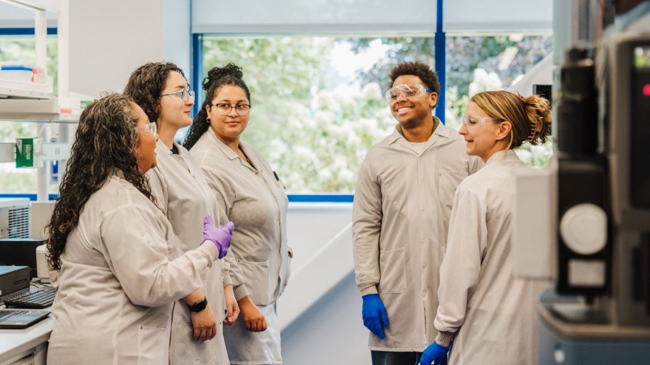 Five smiling Alnylam scientists in lab coats laughing together beside a window in the lab