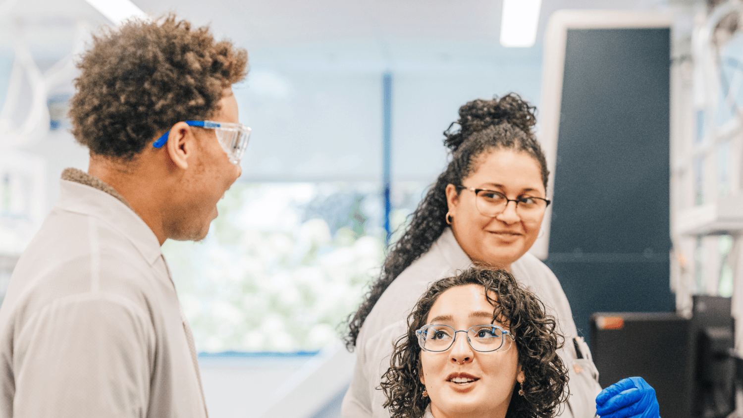 Three Alnylam scientists in lab coats and safety glasses smiling as they talk in the lab