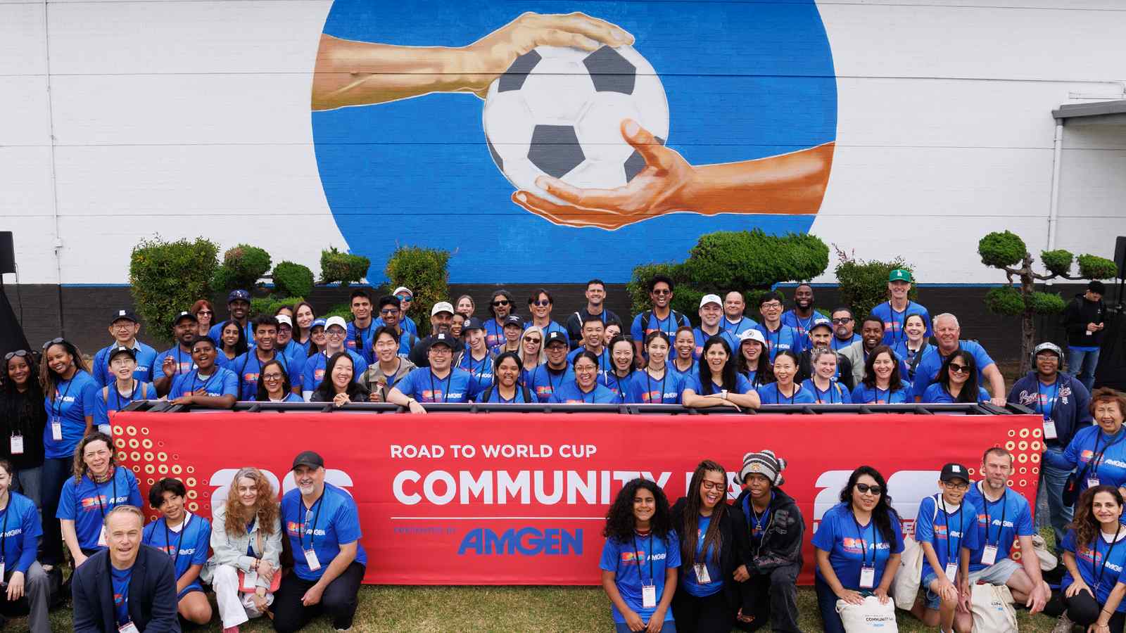 A large crowd of smiling Amgen volunteers in blue shirts gathered behind a Community Day banner