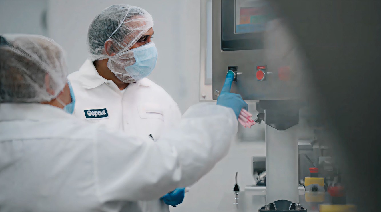 Two Amneal operators in cleanroom whites, hairnets and masks working at a production control panel