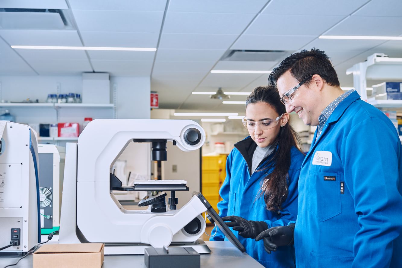 Two Beam scientists in blue lab coats reviewing a tablet beside a microscope in a bright lab