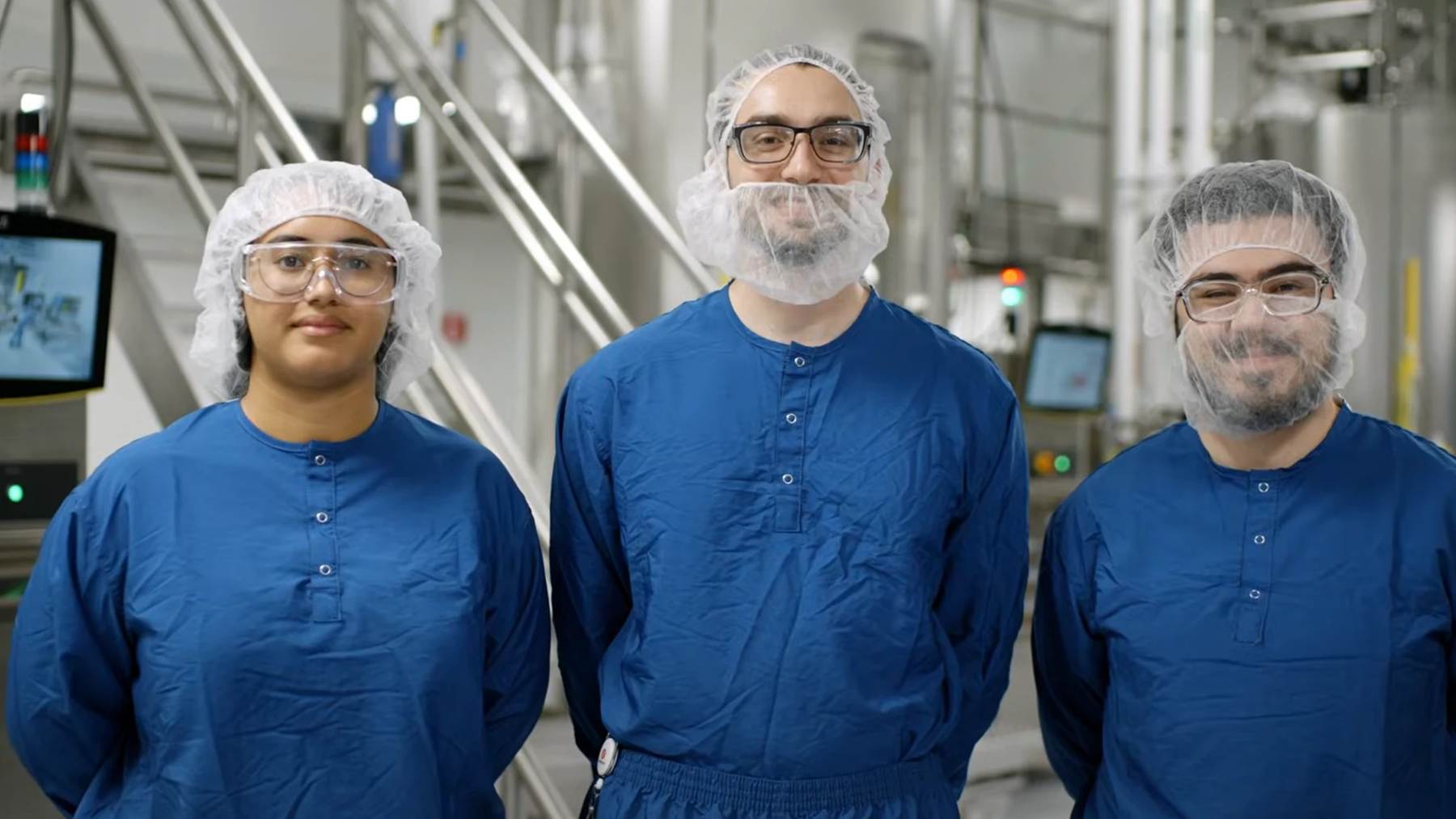 Three smiling BeOne manufacturing colleagues in blue gowns and hairnets standing together on the plant floor