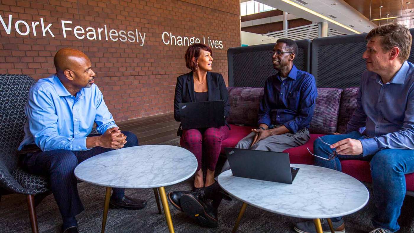 Four Biogen colleagues meeting on sofas beneath a Work Fearlessly, Change Lives wall