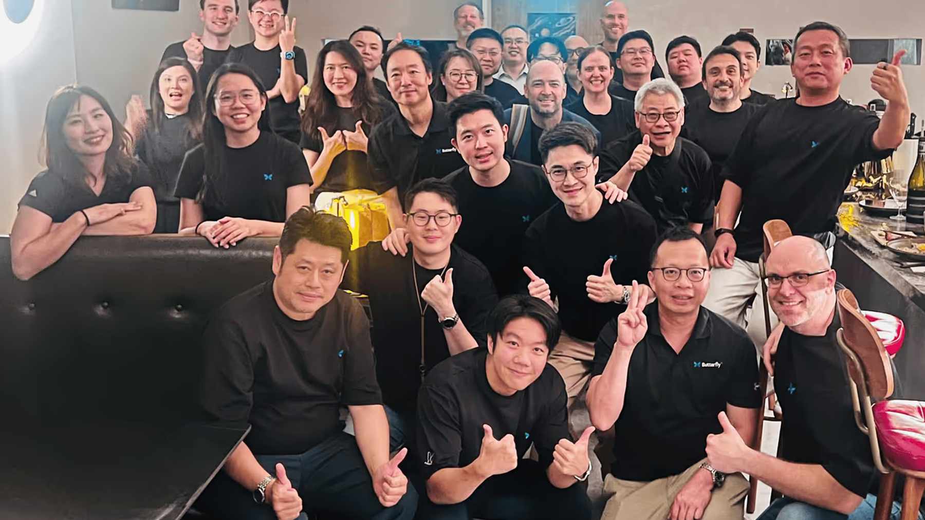 About thirty smiling Butterfly Network colleagues in black Butterfly shirts giving thumbs-up together