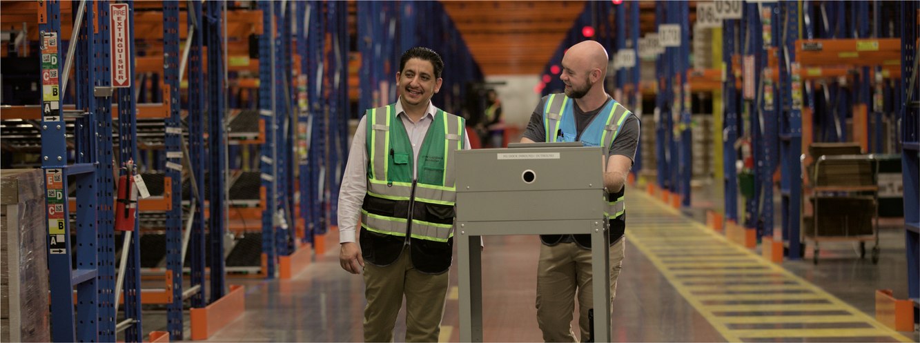 Two Dexcom associates in safety vests walking a cart down a distribution center aisle