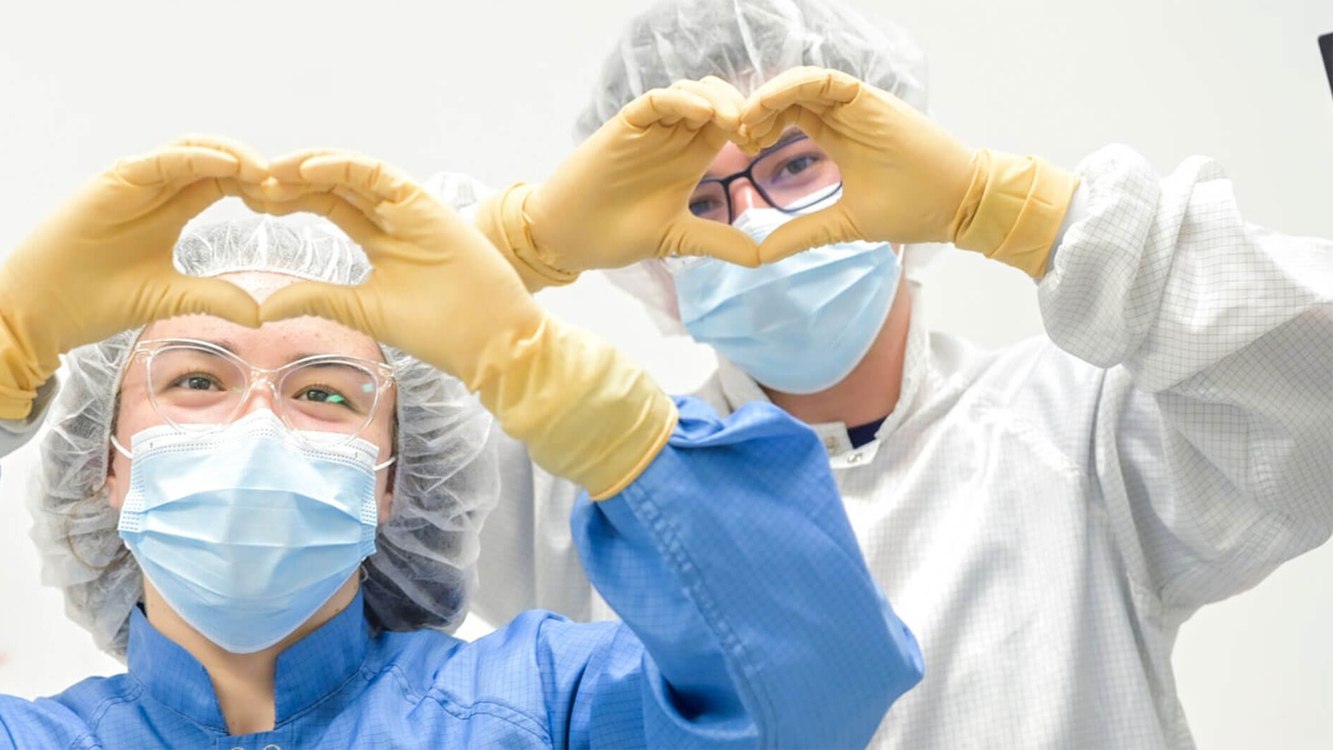 Two Edwards colleagues in cleanroom gowns and masks making heart shapes with their gloved hands