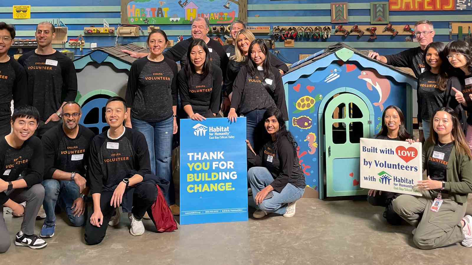 About twenty smiling Exelixis volunteers in matching shirts posing with the playhouses they built for Habitat for Humanity