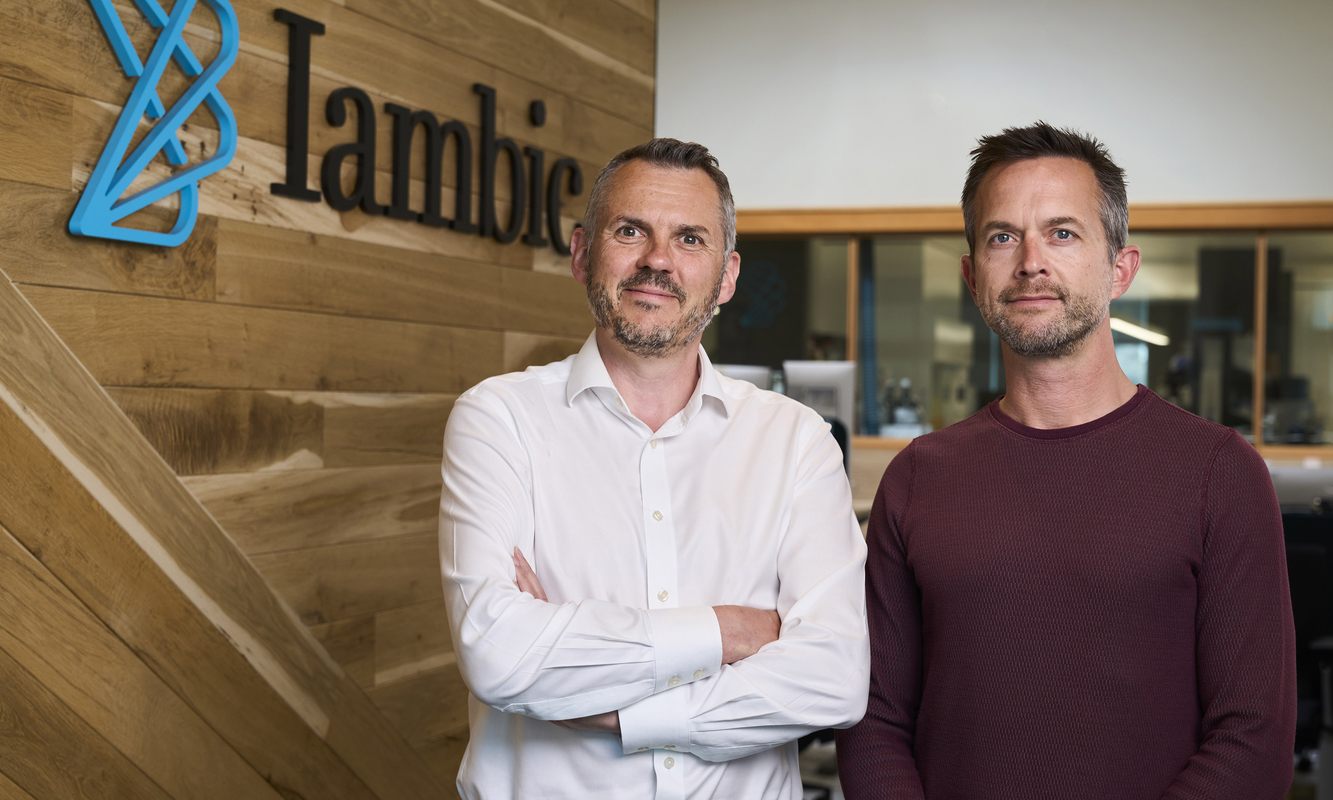 Iambic's two co-founders standing in the office beside the blue Iambic logo on a wood wall