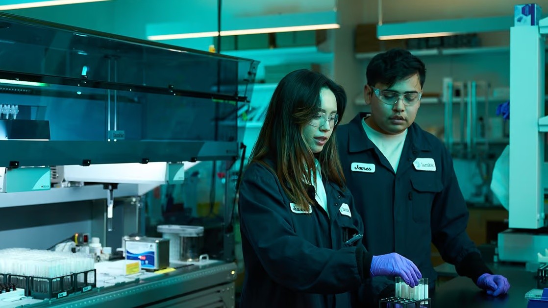 Two Iambic scientists in Iambic lab coats and safety glasses working with sample tubes at a lab bench
