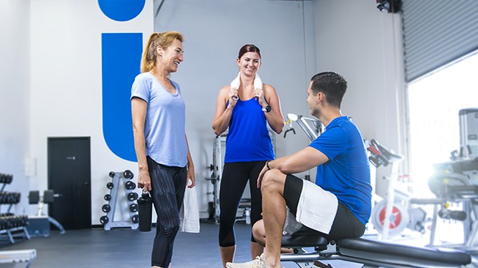 Three smiling ICU Medical colleagues chatting in the company fitness center under a large blue ICU Medical logo
