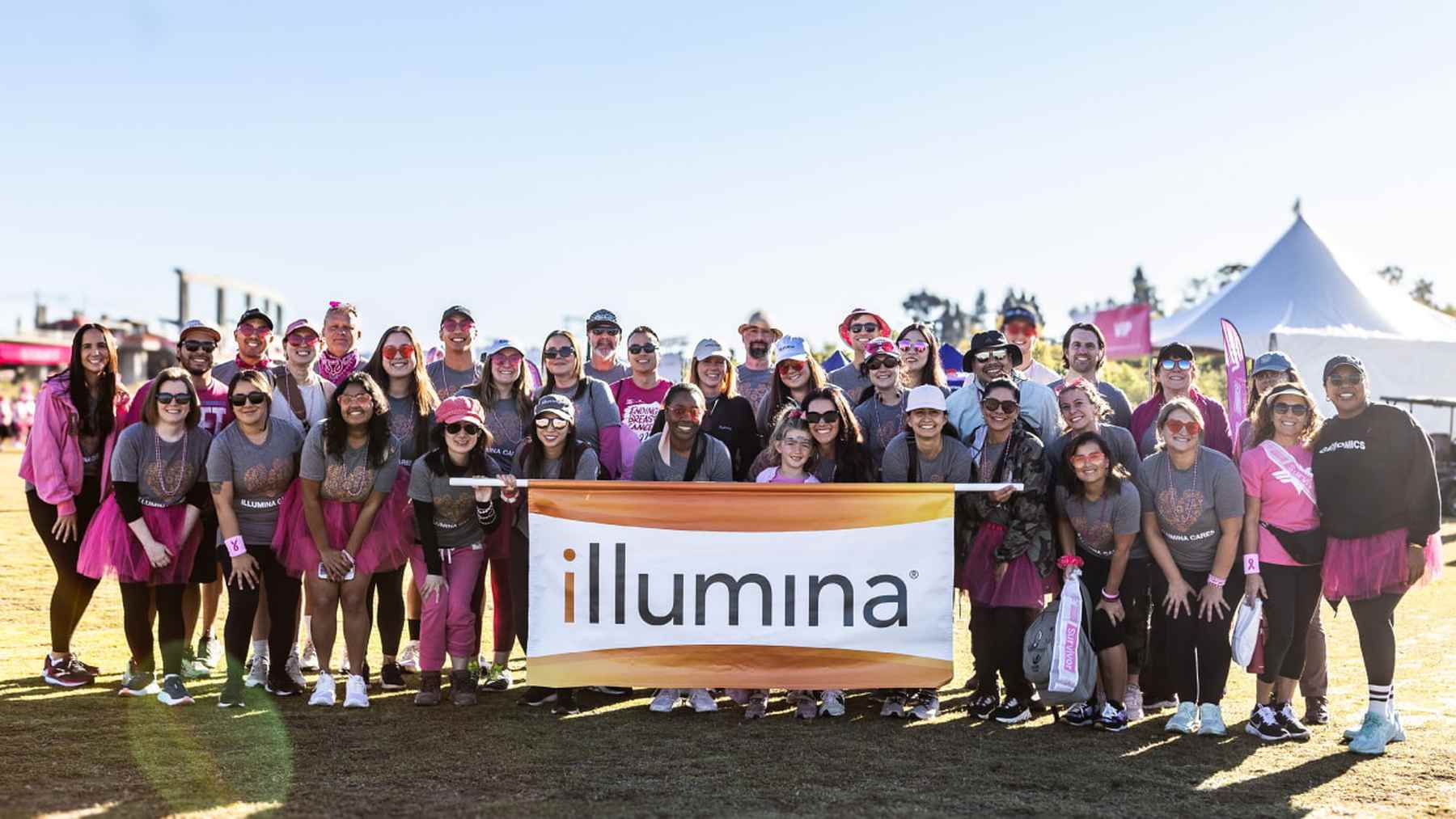 A large team of smiling Illumina colleagues in pink behind an Illumina banner at a charity walk