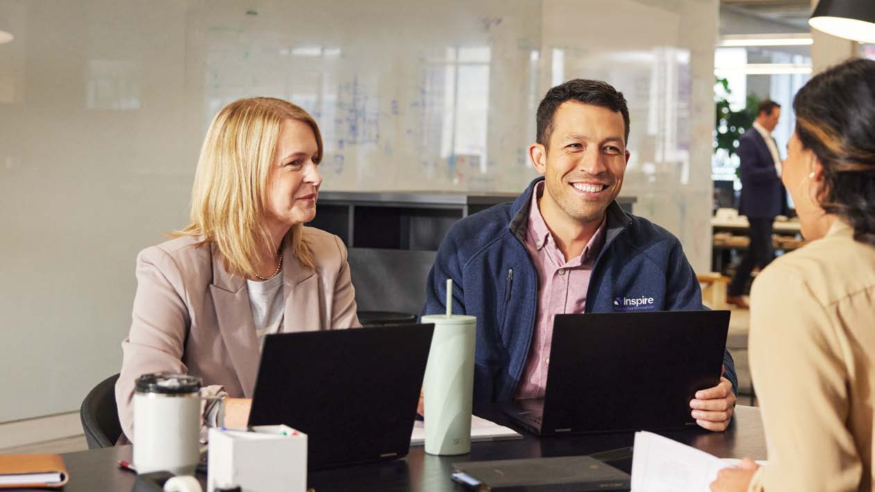 Smiling Inspire colleagues with laptops around a meeting table, one wearing an Inspire fleece