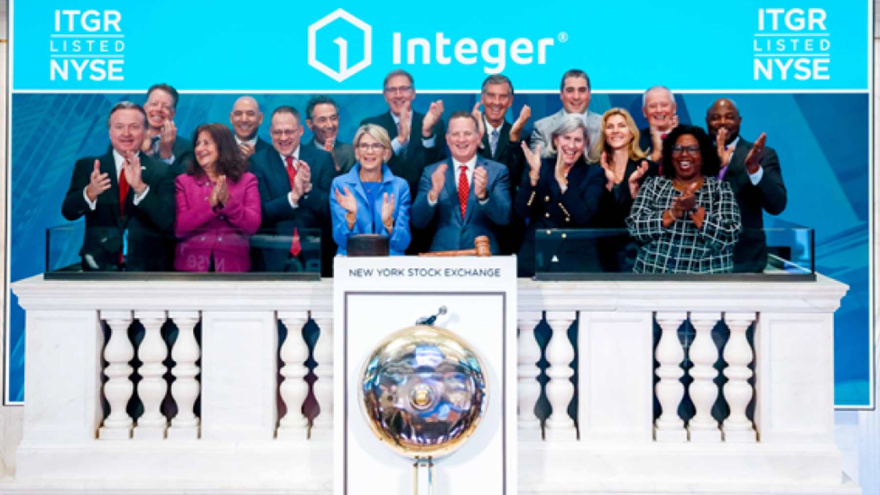 About twenty Integer colleagues clapping together on the New York Stock Exchange bell podium under an Integer banner