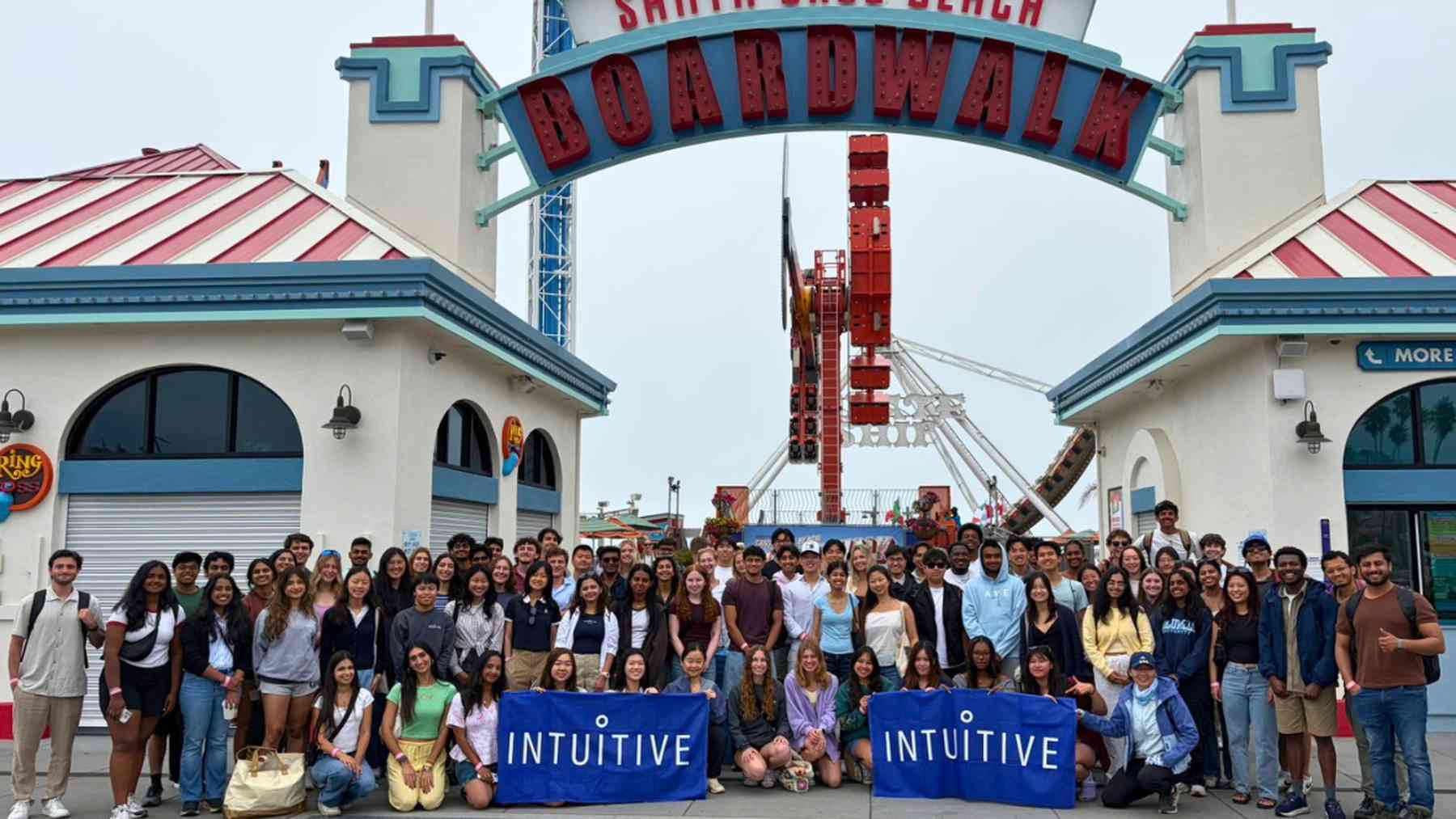 About a hundred Intuitive interns posing together with Intuitive banners under the Santa Cruz Beach Boardwalk arch