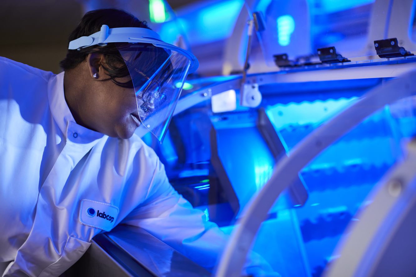 A Labcorp scientist in a face shield working at laboratory equipment lit in blue