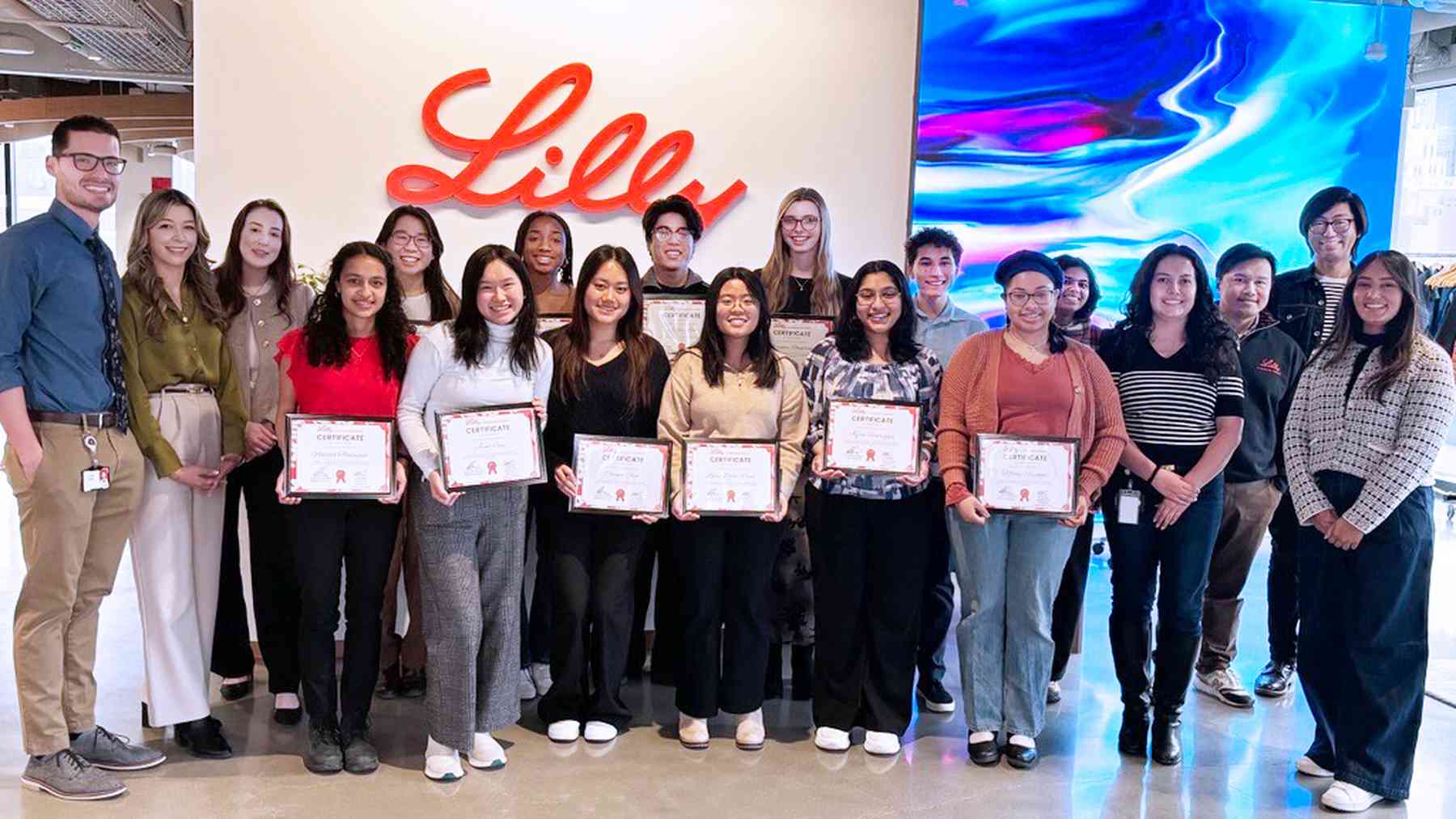About twenty smiling Lilly program participants holding certificates in front of a large red Lilly logo