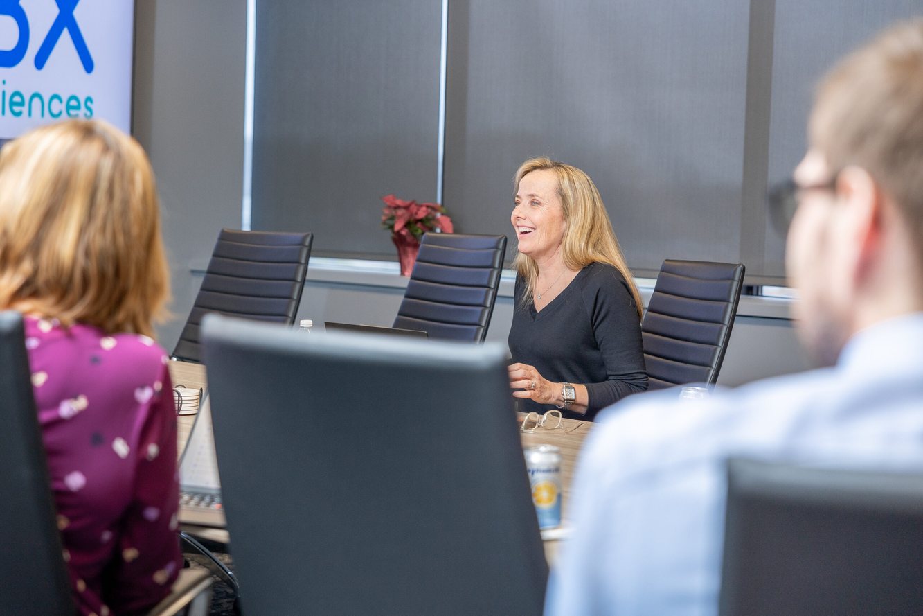 An MBX Biosciences colleague laughing during a team meeting around a conference table, the company logo on the wall