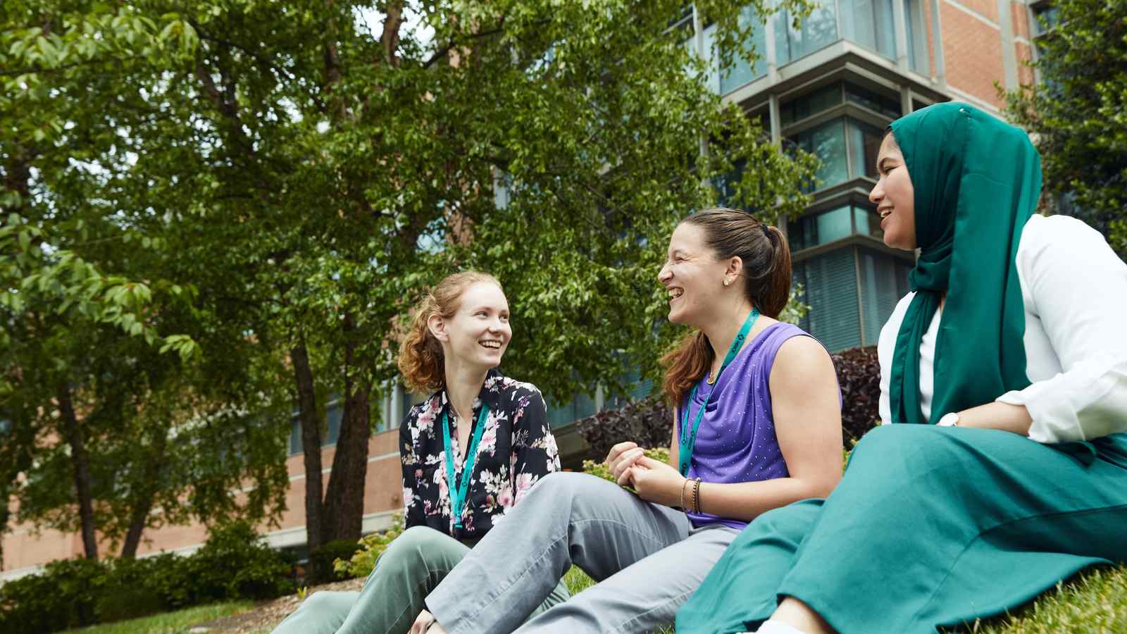 Three Merck colleagues laughing together while sitting on a lawn outside a campus building