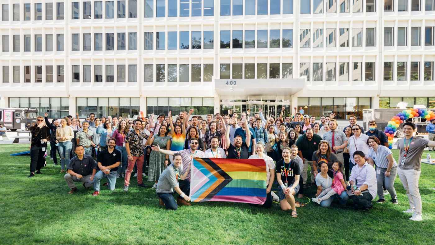 About a hundred smiling Moderna employees gathered on a lawn outside an office building with a Pride flag