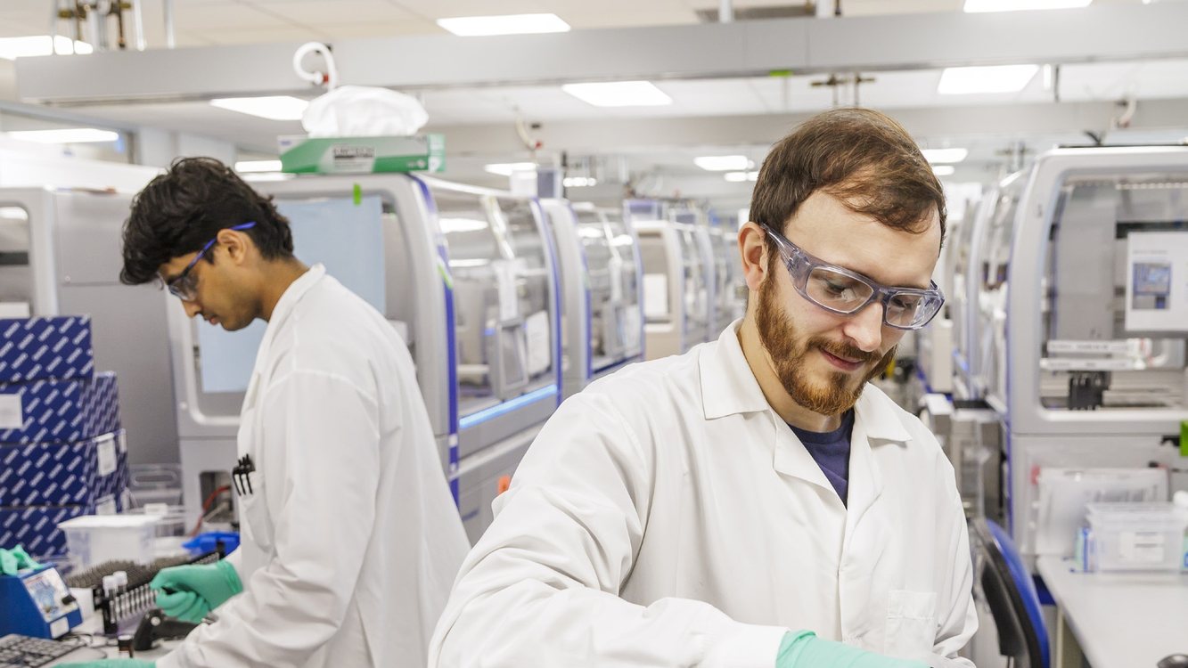 Two Natera scientists in lab coats handling sample plates at a long row of lab instruments
