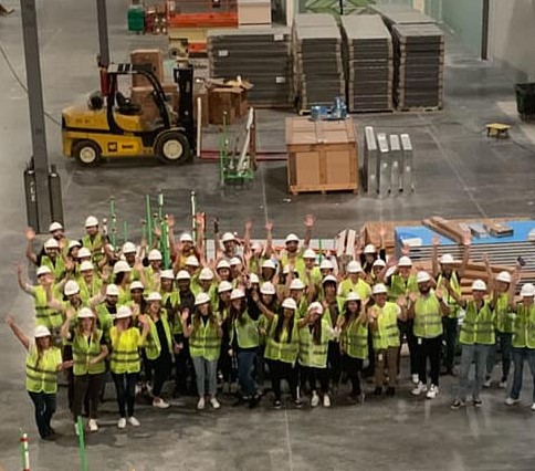 Dozens of Orca Bio colleagues in hard hats and safety vests waving inside a large manufacturing space