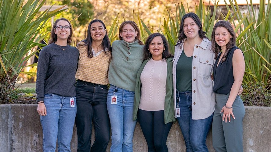 Six smiling Penumbra engineers standing arm in arm outdoors in front of tall plants