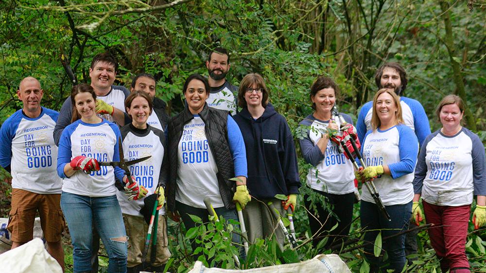 About fifteen smiling Regeneron colleagues in Day for Doing Good shirts holding garden tools in the woods