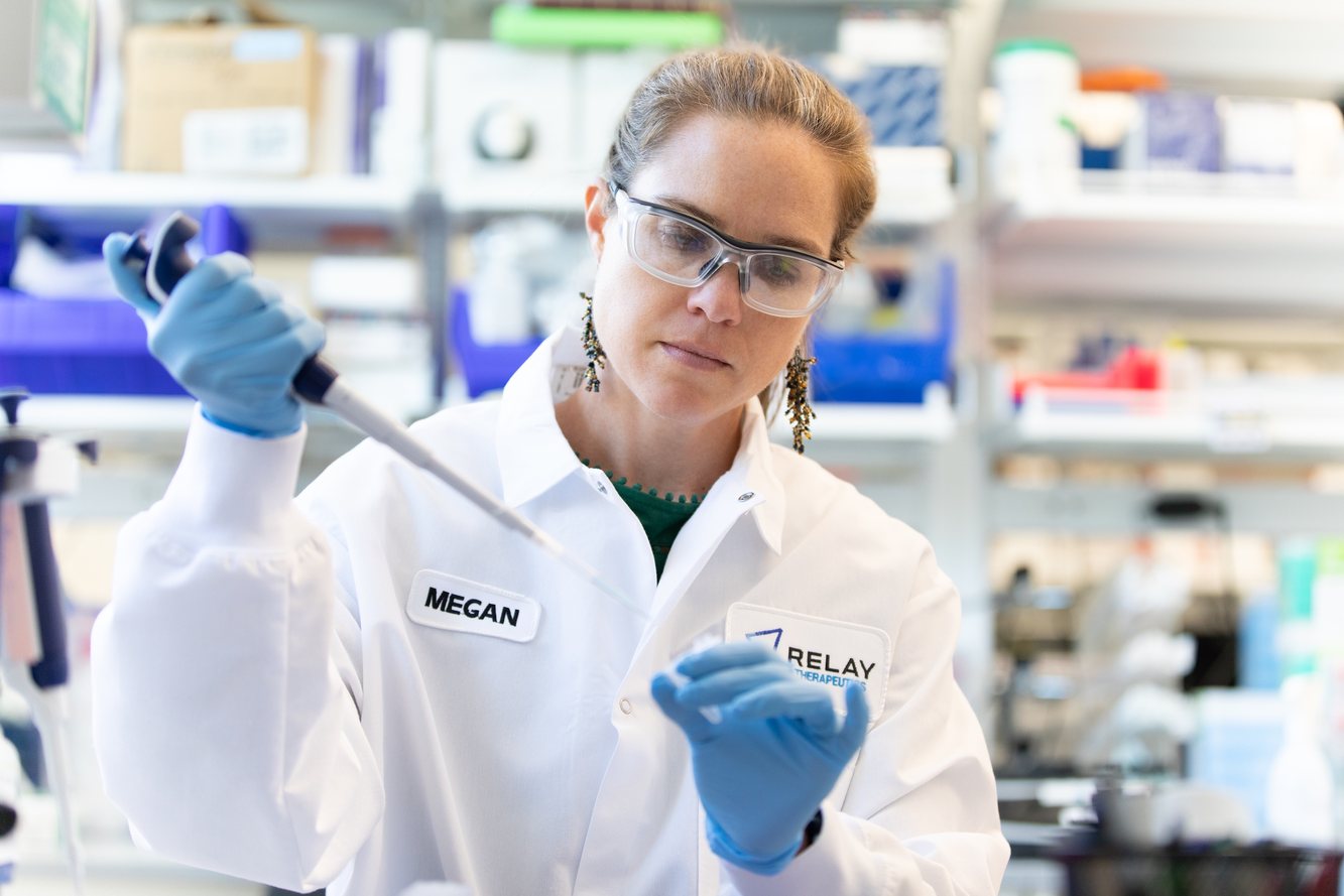 A Relay Therapeutics scientist in a Relay lab coat and safety glasses pipetting at a lab bench