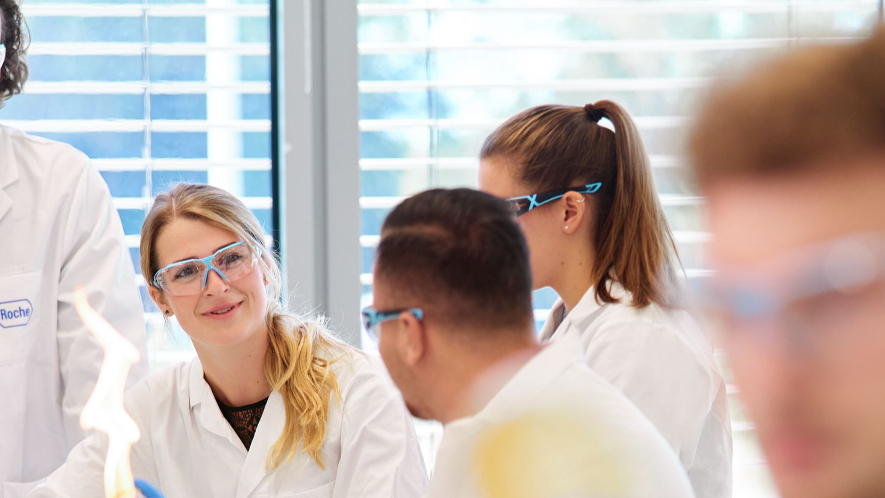 A team of young Roche scientists in lab coats and safety glasses smiling together in a bright lab
