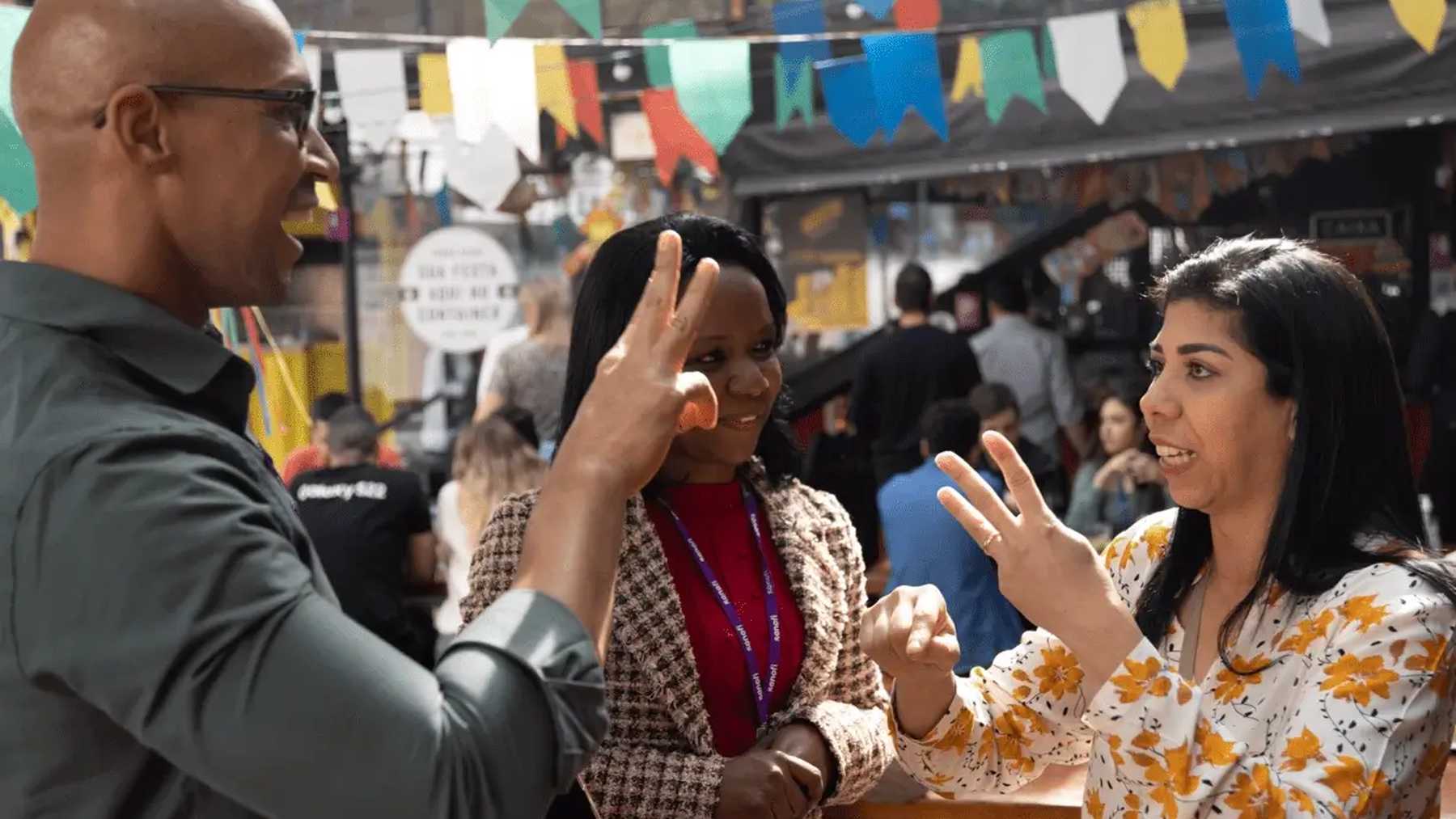 Three smiling Sanofi colleagues talking in sign language at a busy outdoor market