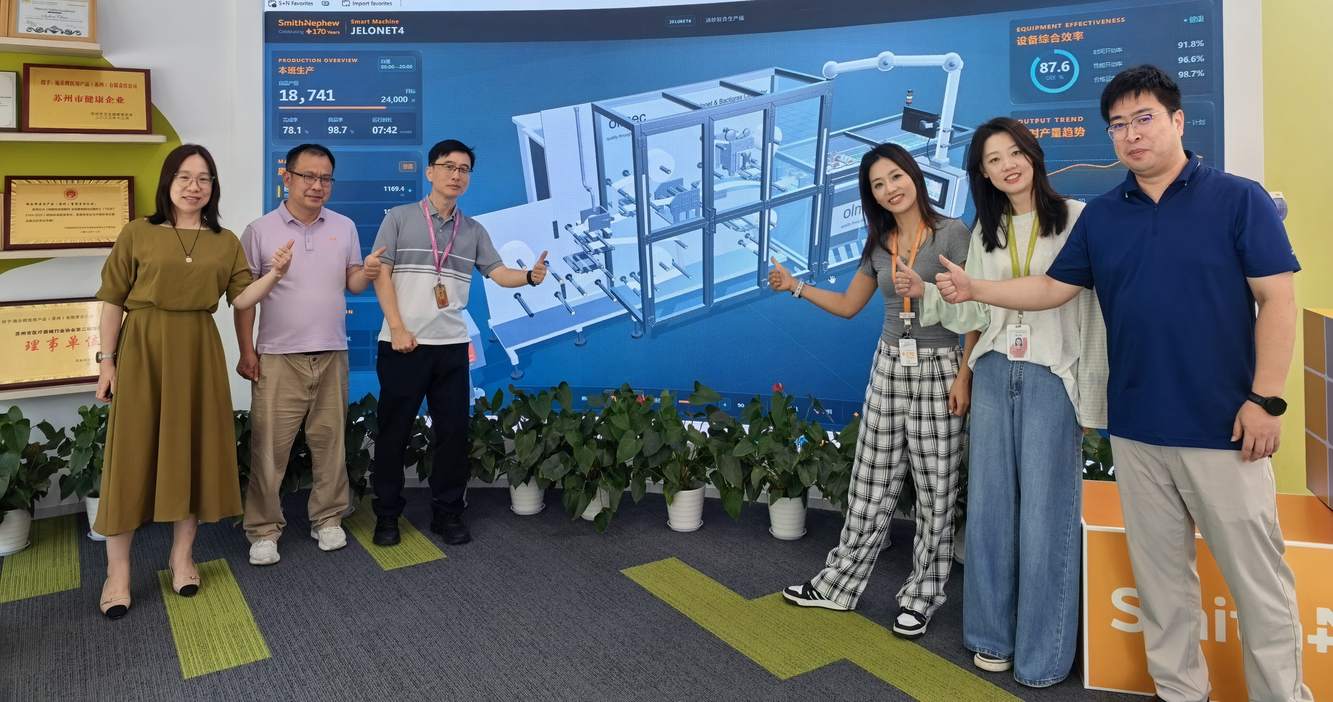 Six Smith+Nephew colleagues, several giving a thumbs-up, in front of a digital production display in the Suzhou factory