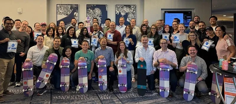 A large group of smiling Spyre colleagues holding skateboards at a company gathering
