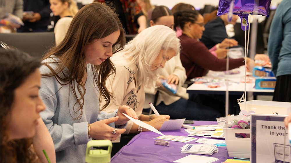 A room full of STERIS colleagues writing cards together at long purple-covered tables during a volunteer event