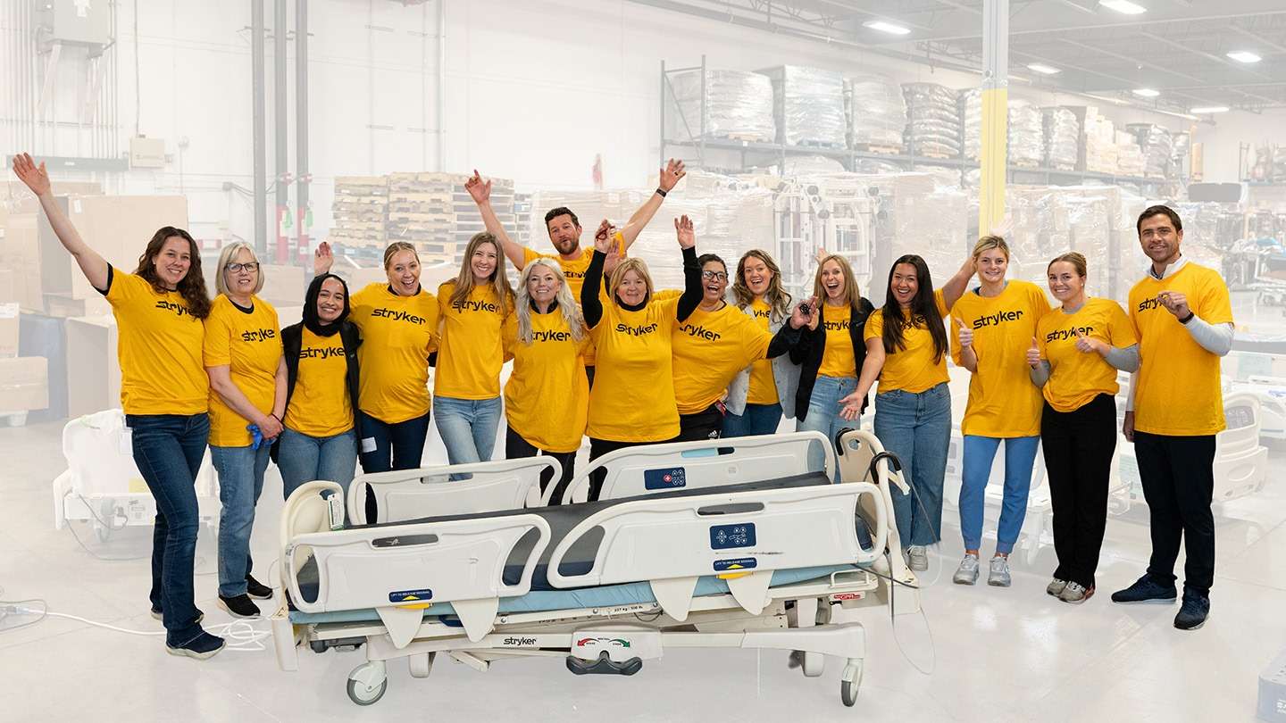 A team of Stryker colleagues in yellow Stryker shirts cheering beside hospital beds in a warehouse