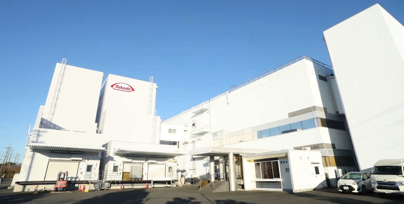 A white Takeda plant building with the red Takeda logo in Narita, Japan, under a clear blue sky