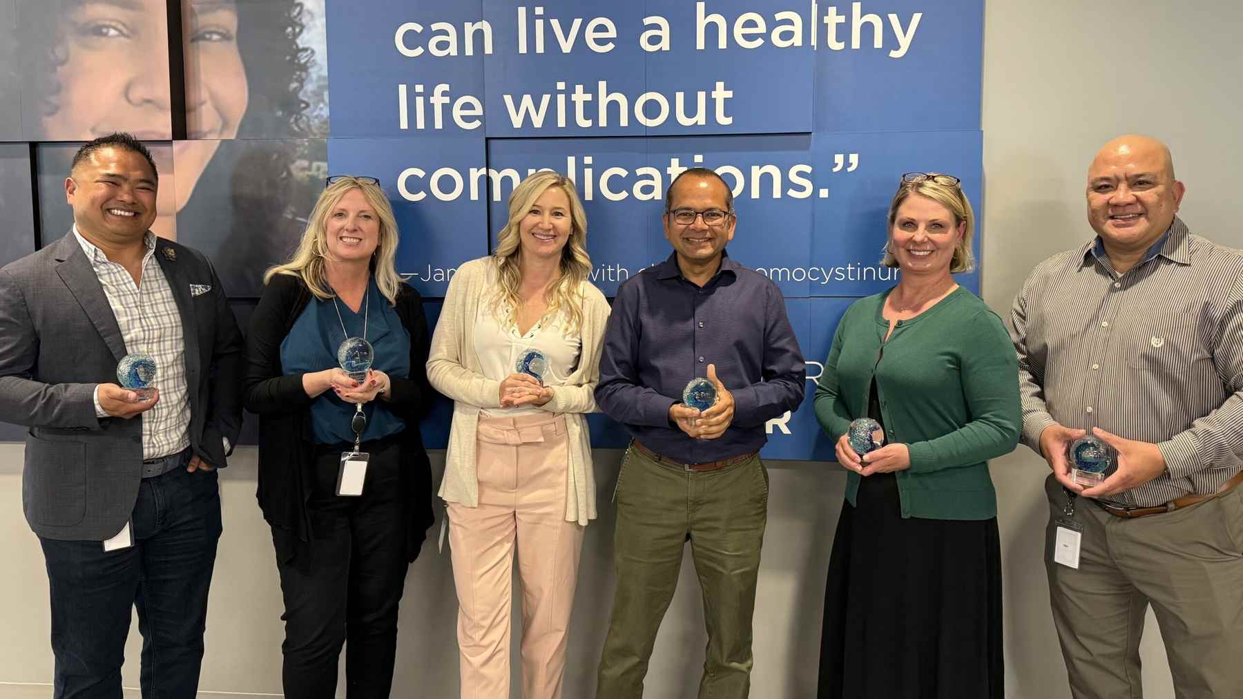Six smiling Travere colleagues holding glass awards in front of a classical homocystinuria banner