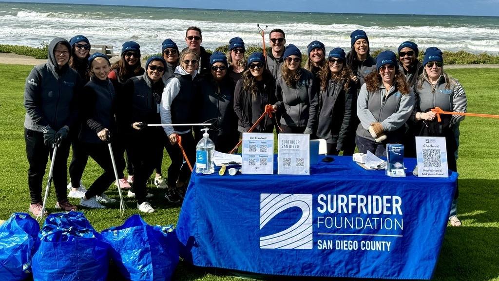 About twenty Travere volunteers in matching beanies at a Surfrider Foundation beach cleanup by the ocean