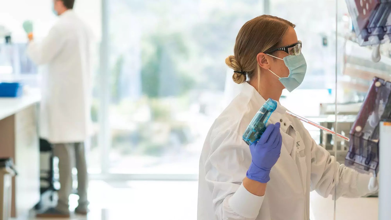 A Vertex scientist in a mask and lab coat working at a lab bench at the San Diego research site